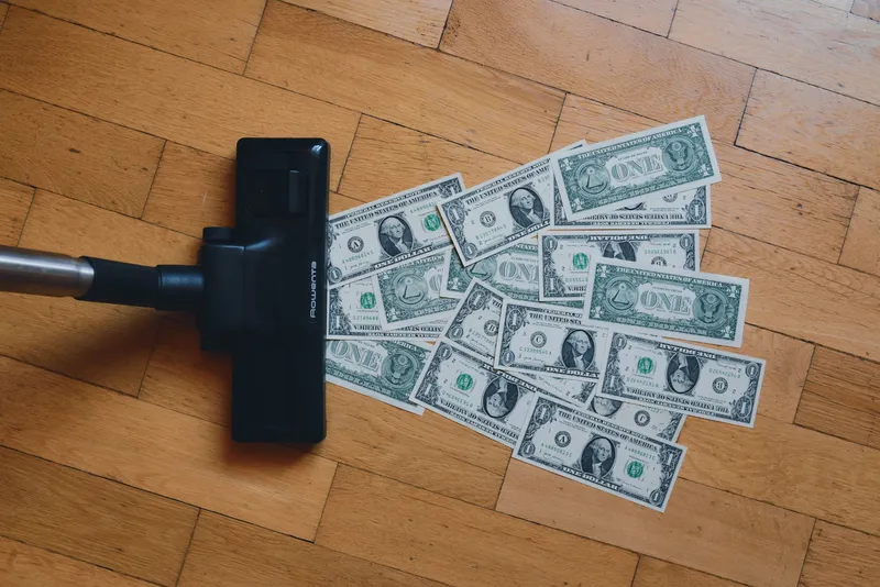 Capul unui aspirator care strânge bancnote de un dolar răspândite pe parchet, simbol al banilor care se pierd