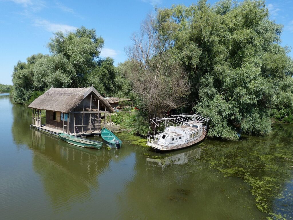 Casă pescărească tradițională cu acoperiș de stuf, pe malul unui canal din Delta Dunării, cu bărci ancorate în față
