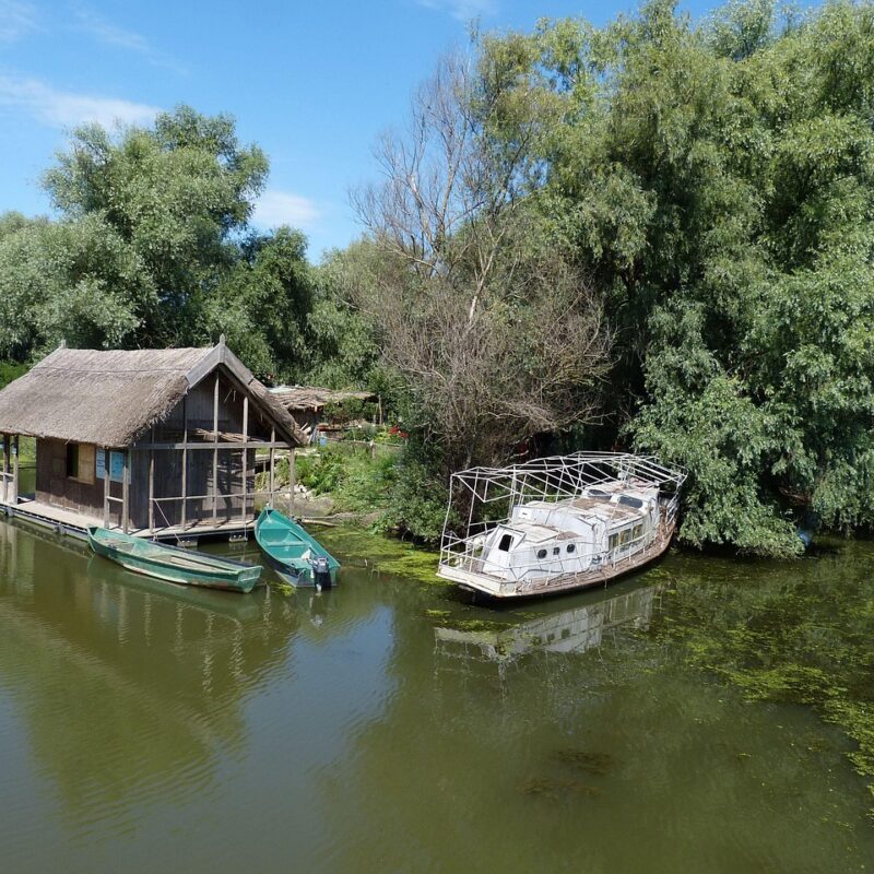 Casă pescărească tradițională cu acoperiș de stuf, pe malul unui canal din Delta Dunării, cu bărci ancorate în față